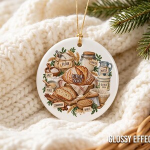 Pu&ograve; includere: Un ornamento rotondo in ceramica bianca con un'illustrazione colorata di ingredienti da forno, tra cui una pagnotta etichettata NAME 2026, farina, lievito madre, acqua, sale, lievito, cannella e grano. Un vasetto reca l'etichetta OLIVIA 2026.