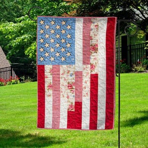 May include: A decorative garden flag featuring a patchwork design of the American flag. The flag has a blue field with wooden stars, red and white stripes, and floral and gingham patterns. The flag is hanging on a black metal stand.
