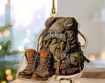 May include: Wooden ornament featuring a backpack, hiking boots, and a belt. The backpack is olive green and brown with the name "John" on it. The boots are brown with laces. A gold string is attached for hanging.