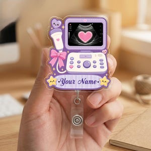 May include: A purple acrylic badge reel featuring a cartoon ultrasound machine with a pink heart on the screen, a matching probe with a pink bow, and a banner at the bottom with the text Your Name flanked by two smiling yellow stars.
