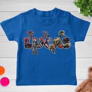 May include: A blue t-shirt featuring the name "LUCAS" in large, colorful letters. The letters are filled with images of Transformers characters. The shirt is made of a soft material and is designed for children.