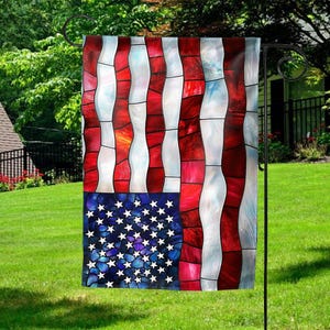 May include: A decorative garden flag featuring a stained-glass design of the American flag. The flag has red and white stripes and a blue field with white stars. The flag is hanging on a black metal stand in a grassy yard.