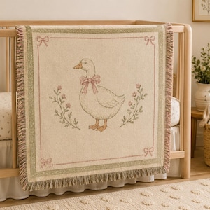 May include: A woven throw blanket featuring a central illustration of a white goose wearing a pink bow, flanked by floral sprigs. The blanket has a cream-colored background, a sage green border, and decorative pink bows in each corner with fringe.