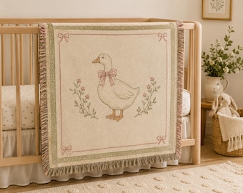 May include: A woven throw blanket featuring a central illustration of a white goose wearing a pink bow, flanked by floral sprigs. The blanket has a cream-coloured background, a sage green border, and decorative pink bows in each corner with fringe.