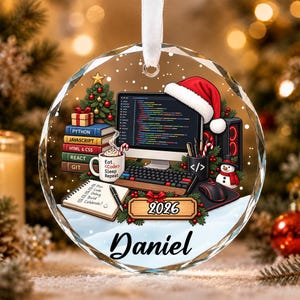 May include: A round glass Christmas ornament featuring a computer setup with a monitor displaying code, a keyboard, a mouse, and a stack of books labeled Python, JavaScript, HTML & CSS, React, and Git. It includes a mug with text, a notepad, and the year 2026 with the name Daniel.