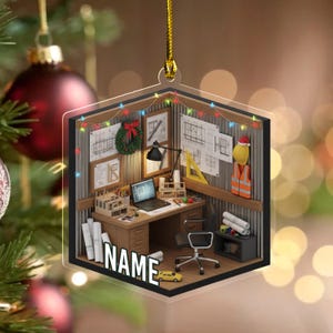 May include: A clear, hexagonal ornament depicting an office scene. The miniature office includes a desk with a laptop, blueprints, a Santa hat, and a construction vest. The word "NAME" is visible on the front.