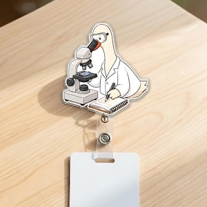 May include: An acrylic badge reel featuring a cartoon goose wearing a white lab coat and safety goggles, looking through a microscope while writing in a notebook. The goose has white feathers and an orange beak, positioned above a clear plastic strap.