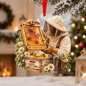 May include: A hanging ornament featuring a beekeeper in a protective suit holding a honeycomb frame with bees. The frame displays the text YOUR NAME and the base shows 202x. It is decorated with white daisies and a metal smoker.