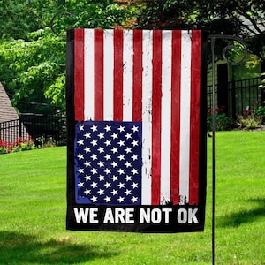 May include: A rectangular garden flag featuring a distressed American flag design. The flag has red and white stripes with a blue square containing white stars. The text "WE ARE NOT OK" is printed in white on a black banner.