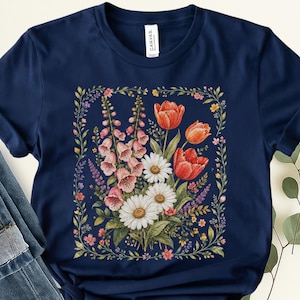 May include: A navy blue t-shirt featuring a central graphic of a floral bouquet with pink foxgloves, white daisies, and orange tulips, all enclosed within a delicate, rectangular vine border with small colorful blossoms and green leaves.