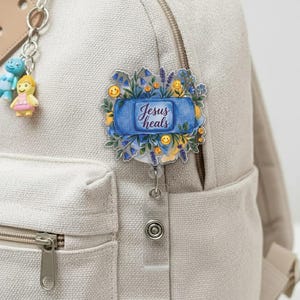 May include: A floral badge reel with the text "Jesus heals" on a blue background. The badge reel is decorated with blue flowers, yellow smiley faces, and lavender sprigs. The badge reel is attached to a beige backpack.