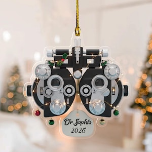 May include: A clear acrylic ornament shaped like an optometrist phoropter, featuring black and silver mechanical details, small holly accents, and tiny hanging ornaments. A white tag at the bottom displays the black text Dr. Sophia 2025.