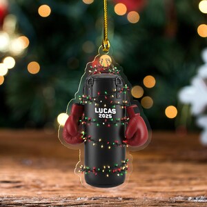 May include: A Christmas ornament shaped like a black punching bag with red boxing gloves. The ornament is decorated with a string of colorful lights and the text "LUCAS 2025". The ornament hangs from a gold string.