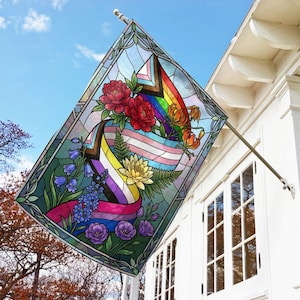 May include: A stained-glass style flag featuring a rainbow, transgender, and non-binary pride design. The flag is adorned with red peonies, orange lilies, bluebells, and purple roses. The flag is displayed outside on a white house with a blue sky background.