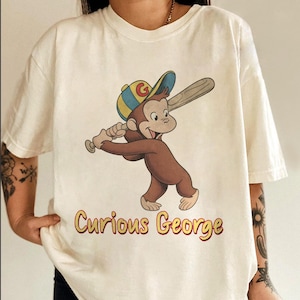 May include: Cream-colored t-shirt featuring a cartoon illustration of Curious George swinging a baseball bat. The monkey wears a baseball cap and the text "Curious George" is printed in yellow and red.