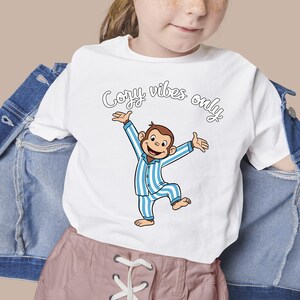 Op de afbeelding: Wit T-shirt met een cartoon aap in blauw en wit gestreepte pyjama, armen uitgestrekt. De tekst "Cozy vibes only" staat in een scriptletter boven de aap. Het T-shirt wordt door een persoon gedragen.
