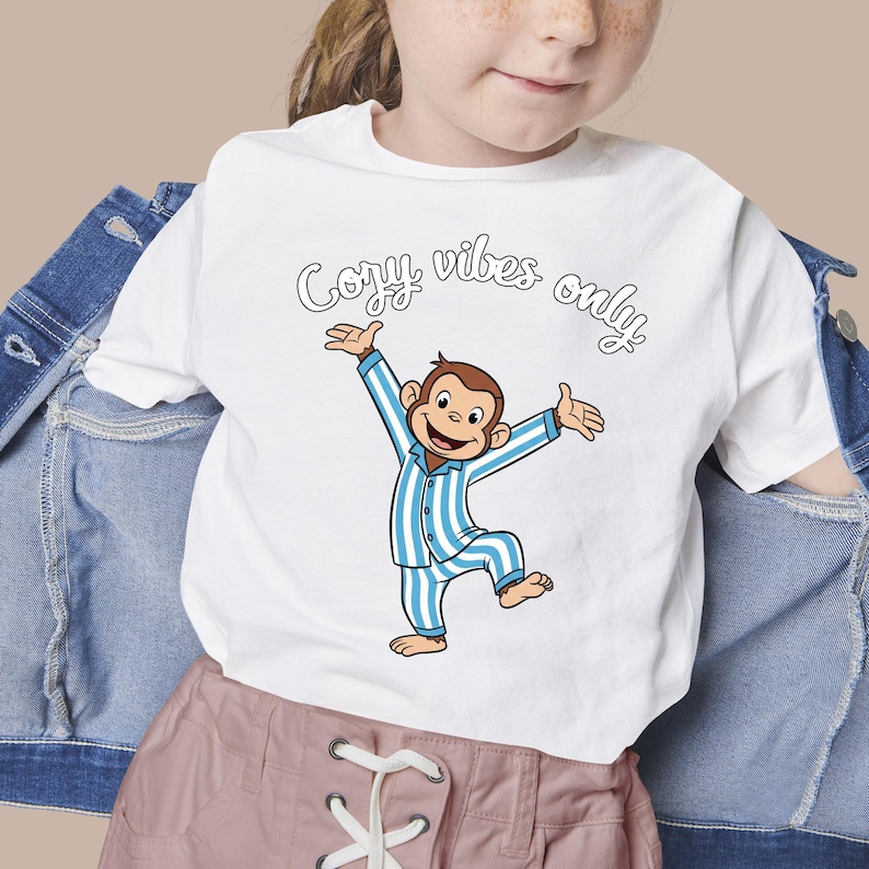Op de afbeelding: Wit T-shirt met een cartoon aap in blauw en wit gestreepte pyjama, armen uitgestrekt. De tekst "Cozy vibes only" staat in een scriptletter boven de aap. Het T-shirt wordt door een persoon gedragen.