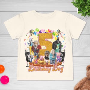 May include: Cream-colored t-shirt featuring a glittery gold number 5, surrounded by cartoon characters and balloons. The words "Birthday Boy" are written in gold script below the characters. A fun, festive design for a child's birthday.