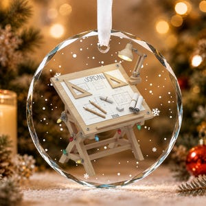 May include: A round, clear glass ornament featuring a wooden drafting table with architectural blueprints, two triangles, pencils, and a desk lamp. The name JORDAN and the year 2026 are printed on the paper. A white ribbon hangs from the top.