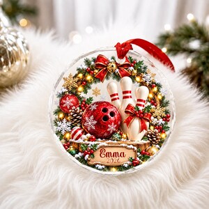 May include: A round, clear Christmas ornament featuring a festive wreath with a red bowling ball, three white bowling pins, pinecones, and red berries. A wooden sign at the bottom displays the name Emma and the year 2026. A red ribbon hangs from the top.