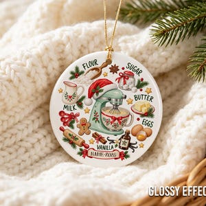 Pu&ograve; includere: Un ornamento rotondo in ceramica bianca a tema pasticceria festiva, con una planetaria verde menta che indossa un cappello da Babbo Natale, circondata da illustrazioni di farina, zucchero, latte, burro, uova, vaniglia e un omino di pan di zenzero.