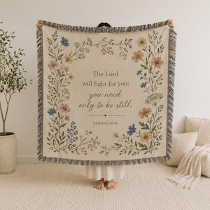 May include: A cream-colored woven throw blanket featuring a floral border with blue, pink, and yellow wildflowers. The center text reads: The Lord will fight for you; you need only to be still. EXODUS 14:14. The blanket has a fringed edge.