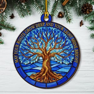 May include: Round wooden ornament featuring a stained-glass tree design in shades of blue and brown. The ornament has the text "MAY YOUR ROOTS GROW DEEP AND YOUR LIGHT SHINE BRIGHT" around the edge. A gold string is attached for hanging.