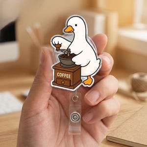 Puede incluir: Un portatarjetas con un dise&ntilde;o de pato blanco con pico y patas naranjas, moliendo granos de caf&eacute; en un molinillo de madera marr&oacute;n con la palabra COFFEE impresa al frente. El dise&ntilde;o es de estilo caricatura con un acabado acr&iacute;lico transparente y brillante.