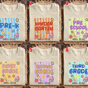 May include: Beige t-shirts with colorful school-themed designs. Each shirt features a grade level: Pre-K, Kindergarten, Preschool, First Grade, Second Grade, and Third Grade. Designs include pencils, apples, backpacks, and stars.