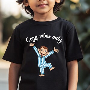 Op de afbeelding: Zwart T-shirt met een cartoon aap in blauw-witte gestreepte pyjama met uitgestrekte armen. De tekst "Cozy vibes only" staat boven de aap. Het shirt is waarschijnlijk voor kinderen.