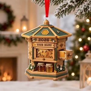 May include: A wooden beehive-shaped Christmas ornament featuring a green roof, the year 2026, and a customizable name plate. The structure includes a honeycomb display, a small pull-out drawer, and two glass jars of honey on a lower shelf.