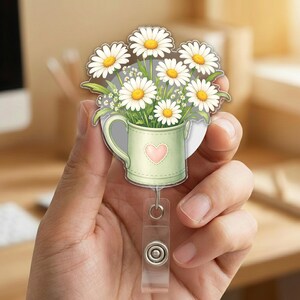 May include: A daisy flower bouquet in a green watering can design on a retractable badge reel. The watering can has a pink heart on the front. The daisies have white petals and yellow centers. The badge reel has a clear plastic strap and a metal clip.
