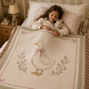 May include: A cream-colored woven throw blanket featuring a central illustration of a goose wearing a pink bow, surrounded by floral sprigs and a decorative border with small bows at the corners, finished with fringed edges on the sides.