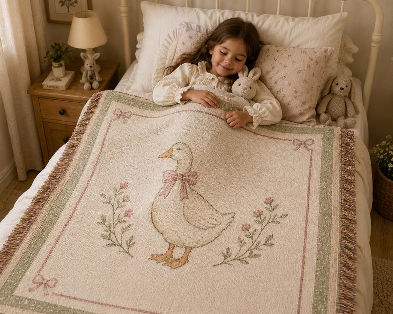 May include: A cream-colored woven throw blanket featuring a central illustration of a goose wearing a pink bow, surrounded by floral sprigs and a decorative border with small bows at the corners, finished with fringed edges on the sides.