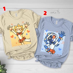 May include: Two t-shirts with cartoon character designs. One is beige with a sun character, the other is light blue with a moon character. Both characters have orange accents and are smiling with their arms raised.