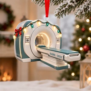 May include: A teal and white MRI scanner Christmas ornament hanging from a red ribbon. The scanner is decorated with a string of colorful holiday lights and a red bow. The side of the scanner bed features the printed text NAME 202X.