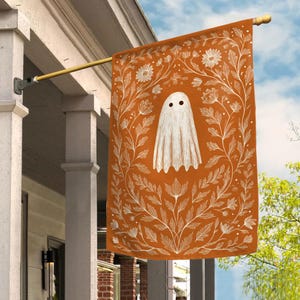 May include: A rectangular garden flag featuring a burnt orange background with a central white ghost illustration. The ghost is surrounded by a detailed white line-art border of various flowers, leaves, and stems, hanging from a gold metal pole.