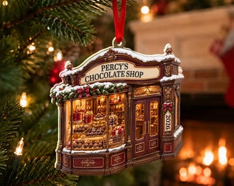 Può includere: Un addobbo per albero di Natale a forma di vetrina con la scritta PERCY'S CHOCOLATE SHOP. L'edificio marrone e rosso presenta un tetto innevato, illuminazione interna, espositori di cioccolatini e l'anno 2026.