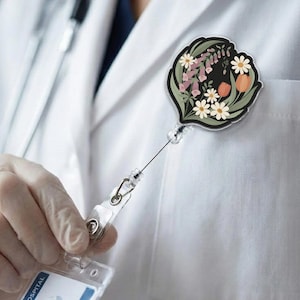 May include: A floral badge reel with a black background and a variety of flowers in shades of pink, white, and orange. The reel is attached to a clear plastic holder and a retractable cord. The badge reel is pinned to a white lab coat.