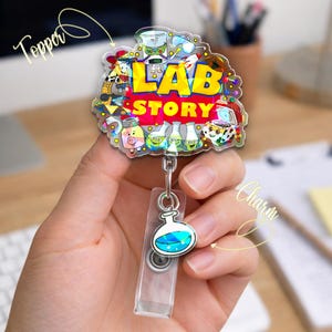 May include: A colorful badge reel with a science theme. The badge reel features the words "LAB STORY" in yellow and red, surrounded by cartoon characters and lab equipment. A clear plastic badge holder and a charm with a blue liquid-filled flask are attached.