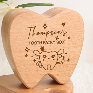 May include: Wooden tooth fairy box with the text "Thompson's TOOTH FAIRY BOX" and a tooth fairy graphic. The box is open, revealing a compartment for baby teeth. The box is made of light-colored wood.