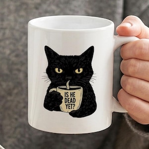 May include: A white ceramic mug featuring a graphic illustration of a black cat with yellow eyes holding a smaller mug. The smaller mug has the text IS HE DEAD YET? printed on it in black capital letters. The mug has a handle on the right side.