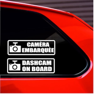 Può includere: Due adesivi rettangolari bianchi con testo nero e icone di fotocamere. L'adesivo superiore recita "CAMÉRA EMBARQUÉE" e quello inferiore "DASHCAM ON BOARD". Gli adesivi sono su un finestrino di un'auto con esterno rosso.
