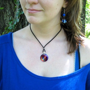 May include: A woman wearing a blue shirt with a black choker necklace and a rainbow swirl pendant. She is also wearing blue and white earrings.