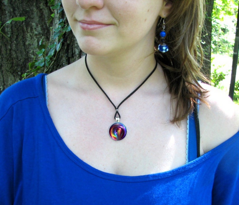 May include: A woman wearing a blue shirt with a black choker necklace and a rainbow swirl pendant. She is also wearing blue and white earrings.