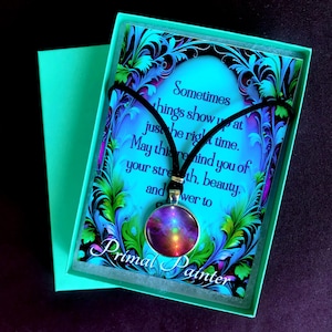 May include: A round pendant necklace with a black cord featuring a colorful abstract design in shades of purple, orange, and yellow. It is displayed on a card with blue and green floral borders and the text "Sometimes things show up at just the right time. May this remind you of your strength, beauty, and power to grow. Primal Painter."