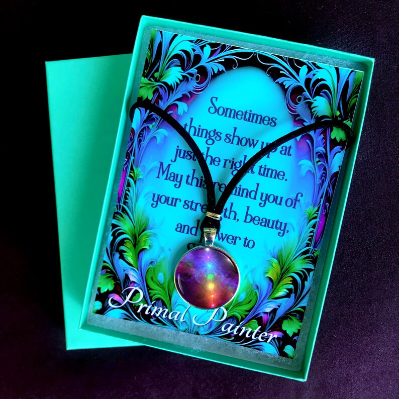 May include: A round pendant necklace with a black cord featuring a colorful abstract design in shades of purple, orange, and yellow. It is displayed on a card with blue and green floral borders and the text "Sometimes things show up at just the right time. May this remind you of your strength, beauty, and power to grow. Primal Painter."