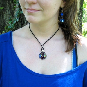 May include: A woman wearing a blue shirt with a black choker necklace and a round pendant with a colorful design. She is also wearing blue and white earrings.