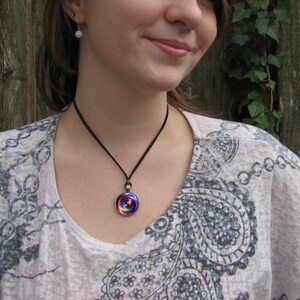May include: A woman wearing a white and gray paisley patterned shirt with a black cord necklace and a colorful iridescent glass pendant.