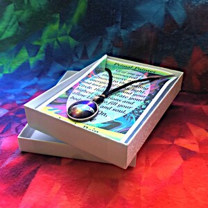 Może przedstawiać: Białe pudełko z czarnym sznurkiem i okrągłym szklanym wisiorkiem z kolorowym obrazem galaktyki w środku. Pudełko zawiera kartkę z tekstem "Primal Painter" i wierszem o uzdrawianiu i świetle.